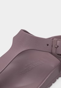 Κοντινό πλάνο λουριού σανδαλιού σε μωβ χρώμα με αγκράφα πάνω σε υφή πέλματος, χαραγμένο με το όνομα της μάρκας.