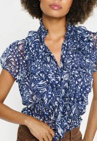 Mornarsko modra bluza z nabranimi robovi z belim paisley vzorcem, prozoren material, kratki rokavi in gumbasta zanka spredaj. Opozarjene nabranosti poudarjajo ovratnik.