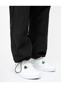 Persona vestida con pantalones negros y zapatillas blancas con acentos verdes, de pie sobre un suelo blanco contra un fondo blanco.
