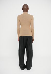 Top cu mâneci lungi, ajustat, de culoare bej, fabricat dintr-un material ușor. Are un detaliu mic cu logo la spate. Este asortat cu pantaloni largi negri.