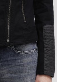 Gros plan d'une personne portant une veste noire avec un patch matelassé sur la manche et une fermeture éclair argentée, associée à un jean bleu en denim.