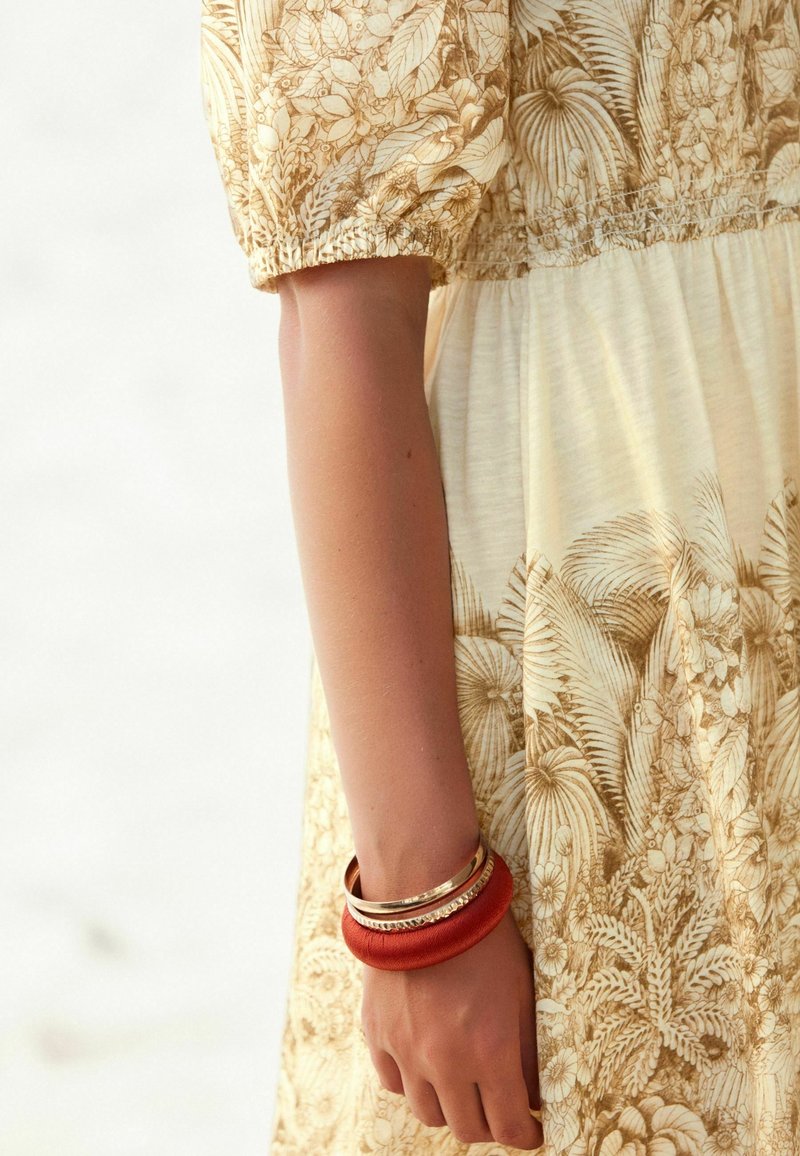 Bras dans une robe fleurie beige à manches courtes bouffantes, portant deux bracelets : un large en cuir rouge et une fine bande dorée.