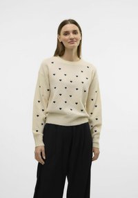 Pull en laine crème avec motif de cœur bleu marine, col rond et poignets côtelés. Associé à un pantalon noir. Fond blanc uni.