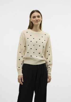 Pull en laine crème avec motif de cœur bleu marine, col rond et poignets côtelés. Associé à un pantalon noir. Fond blanc uni.