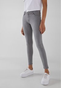 Persona vistiendo jeans ajustados grises, zapatillas blancas y una camiseta blanca metida por dentro, frente a un fondo claro.