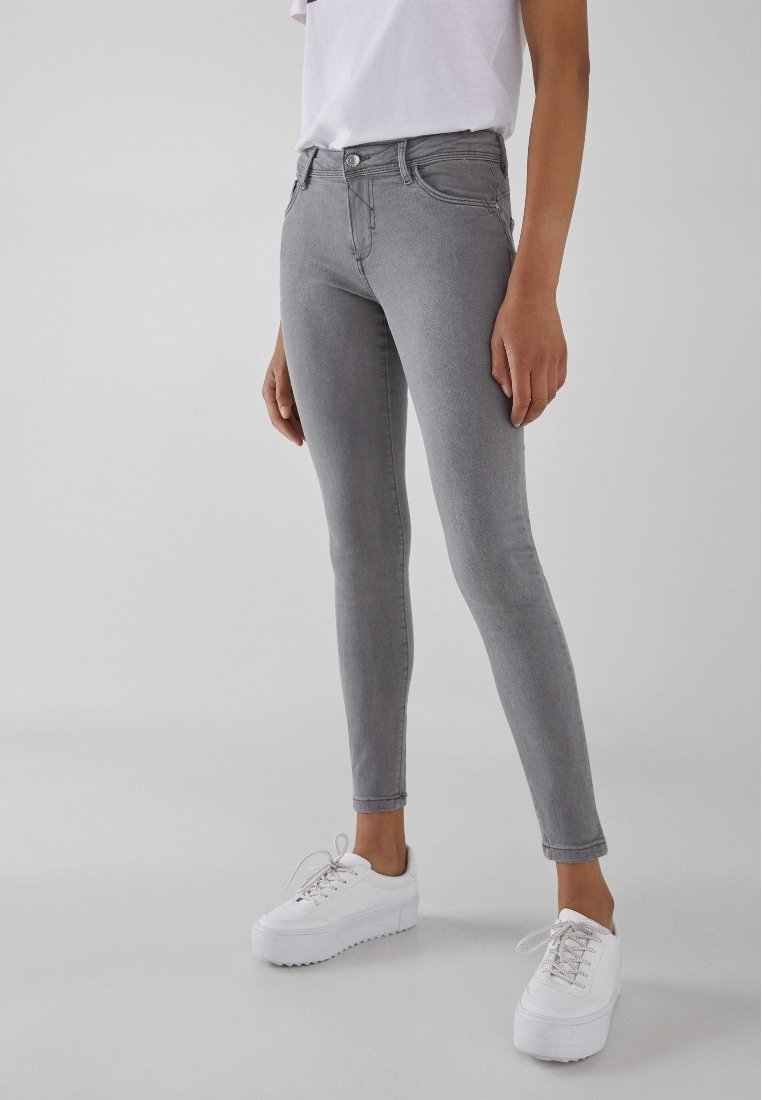 Persona vistiendo jeans ajustados grises, zapatillas blancas y una camiseta blanca metida por dentro, frente a un fondo claro.