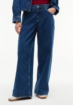 Personne portant un jean bleu à jambes larges à rayures fines, des chaussures blanches et une veste en denim rayée bleue assortie.