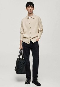 Beige stickad cardigan med knappar, bärs över en vit skjorta, kombinerad med svarta byxor och svarta skor. En svart väska hålls i ena handen.