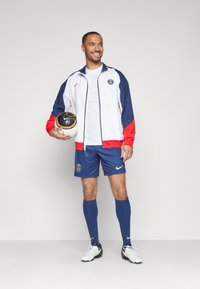 Herrfotbollskläder som består av en vit och röd jacka, blå shorts, blå strumpor och vita fotbollsskor. Inkluderar en svart och vit fotboll.