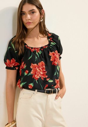 Frau mit glattem braunem Haar trägt eine schwarze Bluse mit rotem Blumenmuster, beige Hose, schwarzen Gürtel und goldene Armbänder, steht vor einer weißen Wand.