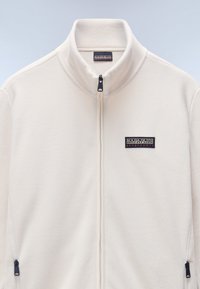Hvid fleece zip-jakke med høj krave, der har et lille sort logo-patch på brystet samt lynlåslommer på siderne.