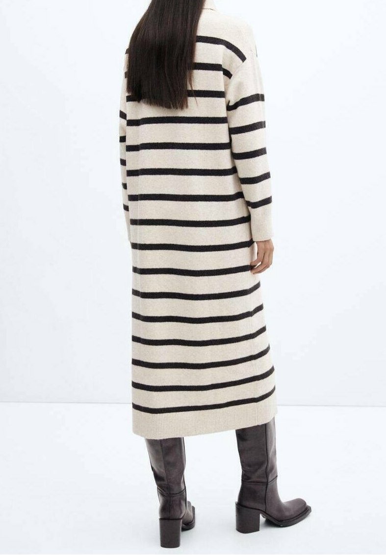 Femme aux longs cheveux foncés portant une longue robe-pull beige et noire à rayures horizontales et des bottes noires montantes à talons, vue de dos.