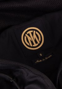 Zwart stoffen interieur met mesh-textuur, met een ronde gouden logo-label en labels die maat (S) en "Gemaakt in Europa" aangeven.