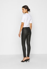 Leggings pretos justos com uma textura suave, cintura alta e detalhes decorativos nas costuras laterais, combinados com uma blusa branca de mangas curtas e saltos pretos.