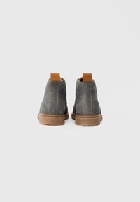 Szare zamszowe botki za kostkę z brązowym skórzanym wzmocnieniem na pięcie oraz teksturowaną gumową podeszwą. Widać detale szycia po bokach.