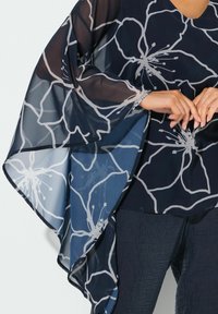 Sheer navy blouse with white floral pattern; features wide, flowing sleeves and a V-neck design; smooth texture, lightweight fabric.