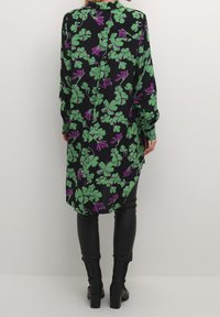 Camisa larga negra con patrón floral verde y morado, con botones en la parte posterior y dobladillo redondeado, combinada con pantalones ajustados negros y botas de tobillo.