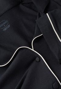 Zwarte pyjamashirt met witte piping langs de kraag en de knopenlijst, met een zachte stofstructuur en een label dat de maat en herkomst aangeeft.