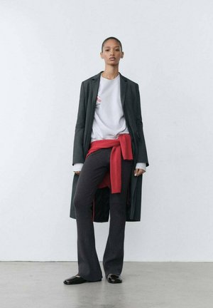 Czarny długi płaszcz noszony na jasnoszarej koszulce, czerwony sweter związany w talii oraz ciemne dzwony. Błyszczące czarne buty. Gładkie tekstury w całym zestawie.