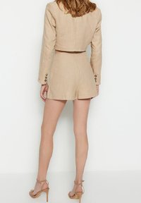 Completo due pezzi in lino beige con giacca corta e pantaloni a vita alta, caratterizzato da bottoni sulle maniche. Texture liscia.