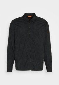Veste noire à manches longues zippée avec col, motif texturé subtil et petit patch logo sur la manche gauche.