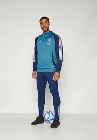 Mannen sportsoutfit met een teal pullover hoodie met marineblauwe accenten en logo, gecombineerd met marineblauwe sportbroeken en zwarte voetbalschoenen, naast een voetbal.