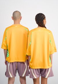Două persoane poartă tricouri mari, supradimensionate, de culoare portocalie cu accente verzi, asortate cu pantaloni scurți gri cu dungi albe și detalii din plasă.