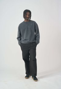 Grauer Sweatshirt mit Rundhalsausschnitt, ausgestattet mit einem kleinen blauen Logo, kombiniert mit schwarzen, locker sitzenden Hosen und dunklen Schuhen.