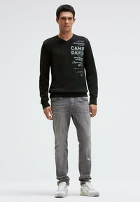Schwarzer V-Ausschnitt-Pullover mit gedrucktem Text, kombiniert mit hellgrauen Jeans mit Used-Details. Gestylt mit weißen Turnschuhen.