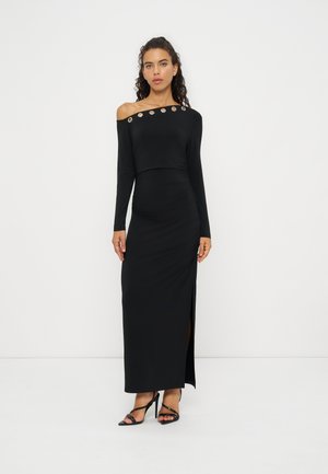 Femme aux cheveux bouclés portant une longue robe noire épaules dénudées avec des détails œillets en métal et des talons hauts noirs à brides, debout devant un fond blanc.