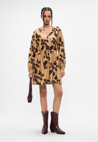 Καφέ και μαύρου χρώματος μοτίβο φόρεμα με μακριά μανίκια και V-τύπου λαιμόκοψη. Διαθέτει σφιχτή μέση και ροή στη φούστα. Συνδυάζεται με καφέ μπότες με μυτερή μύτη.