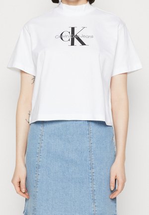 Biały krótki T-shirt z logo "Calvin Klein Jeans" noszony z jasnoniebieską dżinsową spódnicą, osoba stoi z ramionami swobodnie opuszczonymi wzdłuż ciała.