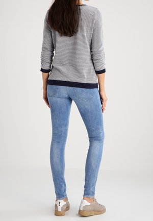 Woman with long hair wearing light blue skinny jeans, gray textured sweater with dark cuffs, and beige sneakers standing facing away.