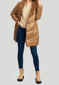 Donkergroene pufferjas met een pluche capuchon, voorzien van drukknoopsluitingen en een geborduurd patroon, gecombineerd met blauwe jeans en beige enkellaarsjes.