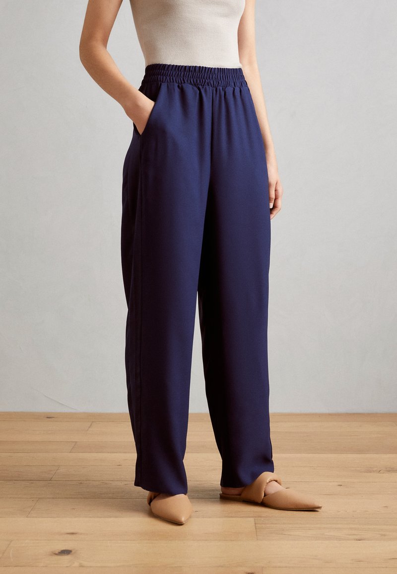 Pantalons bleu marine à coupe ample avec une taille froncée, poches latérales et une texture lisse, assortis à des chaussures à bout pointu beiges.