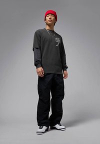 Dunkelgraues kurzärmliges Hemd über einem schwarzen langärmligen Hemd, kombiniert mit schwarzen Cargo-Hosen und weißen Sneakers mit schwarzen Akzenten. Rote Mütze.