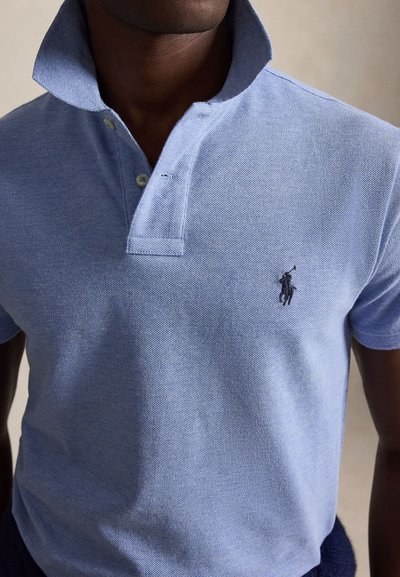 Mężczyzna noszący jasnoniebieską koszulkę polo z podniesionym kołnierzem i czarnym, wyszywanym logo gracza polo na piersi.