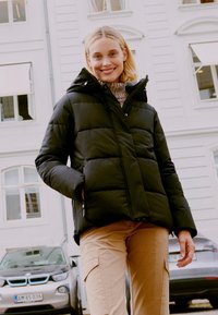 Jachetă puf cu design acoladă, de culoare neagră, cu glugă, având buzunare laterale și fermoar. Purtați-o peste un pulover bej și pantaloni cargo bej.