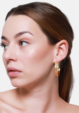 Frau mit zurückgebundenem braunem Haar, schaut nach links, trägt einen goldfarbenen, strukturierten Creolen-Ohrring in Form einer Feder vor einem schlichten weißen Hintergrund.