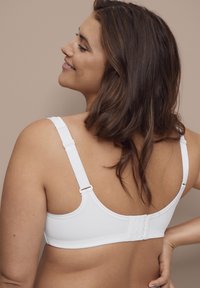Vrouw met half-lang bruin haar, gekleed in een witte beha, glimlachend en kijkend naar de zijkant tegen een neutrale beige achtergrond.