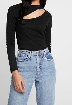 Woman wearing a black long-sleeve top with a diagonal chest cutout and high-waisted light blue jeans, standing against a plain background.