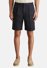 Pantaloni scurți pliați de culoare azul închis, confecționați dintr-un material ușor, cu o croială lejeră și buzunare laterale. Asortați cu pantofi slip-on bej.