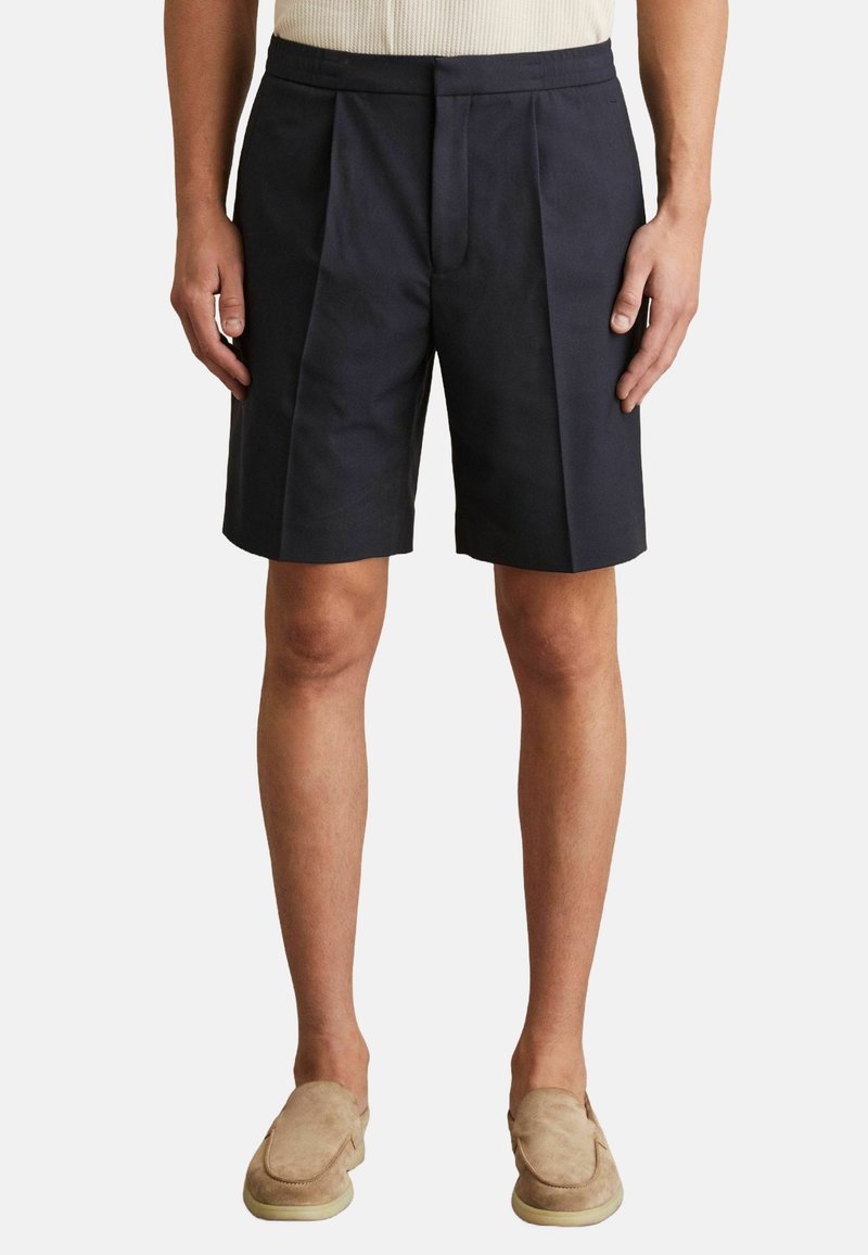Pantaloni scurți pliați de culoare azul închis, confecționați dintr-un material ușor, cu o croială lejeră și buzunare laterale. Asortați cu pantofi slip-on bej.