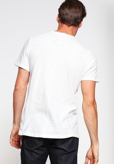 Hvit T-skjorte med korte ermer, teksturert overflate og synlige sømmer, kombinert med mørke jeans, sett fra baksiden.