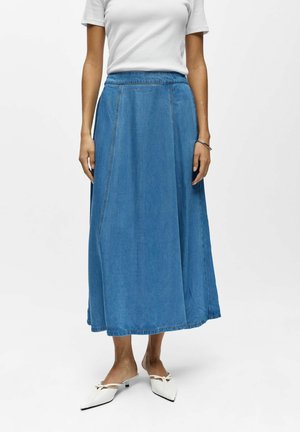 Žena nosi bielu košeľu s krátkym rukávom, modrú džínsovú sukňu po členky a biele špicaté topánky s nízkym podpätkom a mašličkovými detailmi.