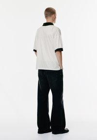 Person st�år med ryggen til iført en let ternet polo-shirt med sort krave og ærmer, løse sorte jeans og sorte sko.