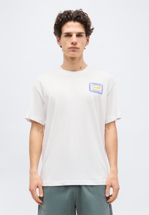 Νέος άνδρας με σγουρά σκούρα μαλλιά, φορώντας ένα λευκό μπλουζάκι με ένα μικρό λογότυπο «Original Sardines» στο στήθος και γκρι σορτς.