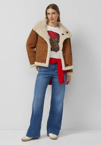 Brązowa kurtka z sztucznego zamszu z kremowym kołnierzem z sztucznego futra, zapinana na guziki, połączona z białym t-shirtem z grafiką, czerwoną paskiem i szerokimi niebieskimi dżinsami.