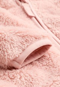 Vêtement en tissu rose duveteux avec une texture douce et pelucheuse, doté d'un ourlet lisse et d'une ouverture à l'extrémité de la manche.