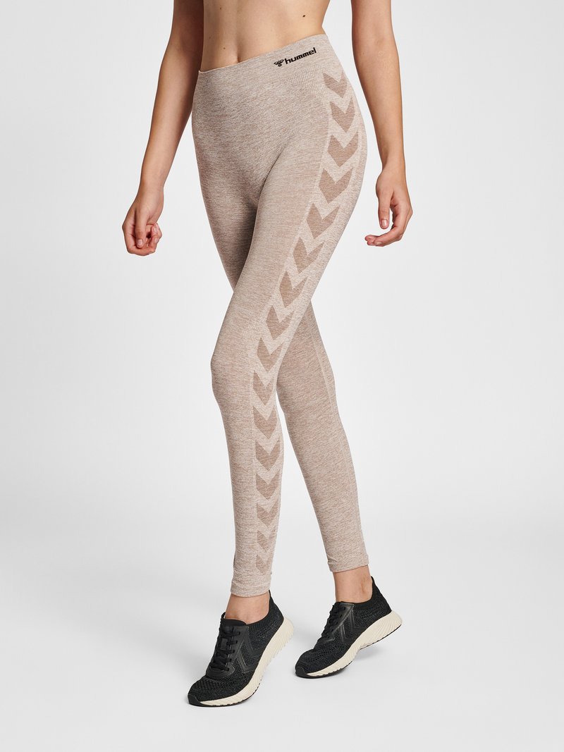 Vrouw met beige, hooggetailleerde legging met chevronpatronen langs de benen en zwarte sportschoenen op een effen achtergrond.
